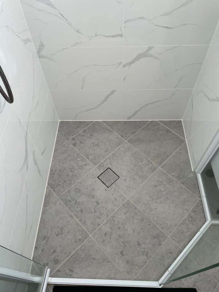 A top-down view of a walk-in shower floor featuring light grey, speckled tiles laid in a diagonal diamond pattern with a central square drain.