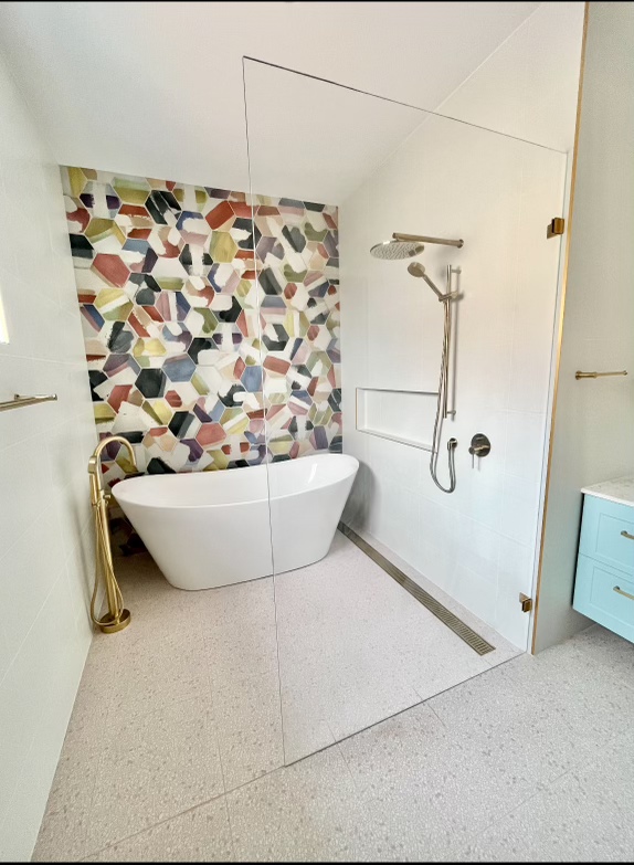 A modern bathroom with a freestanding white bathtub against a colourful mosaic-tiled wall, a glass-enclosed shower area with a rain showerhead, and a light blue vanity