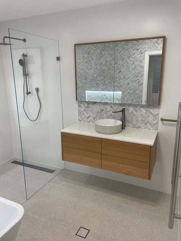 A modern bathroom with a walk-in glass shower, a wooden vanity with a white countertop and a round vessel sink, a large mirror with a herringbone patterned tile backsplash, and grey tiled flooring